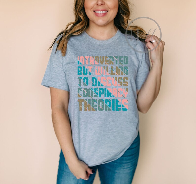 May include: A gray t-shirt with a graphic design that reads "Introverted but willing to discuss conspiracy theories". The text is in a colorful, striped font.