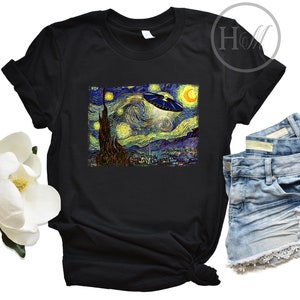 May include: A black t-shirt with a graphic print of Vincent van Gogh's Starry Night with a UFO flying over the town. The t-shirt is laying on a white surface with a pair of blue denim shorts.