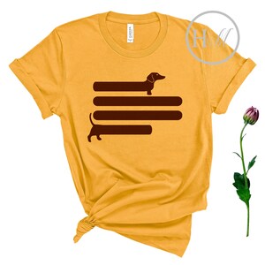 May include: A yellow t-shirt with a brown dachshund silhouette design. The dachshund is depicted walking across four horizontal lines.