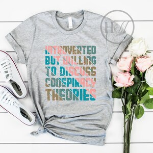 May include: Gray t-shirt with the text "Introverted but willing to discuss conspiracy theories" in a colorful retro font.