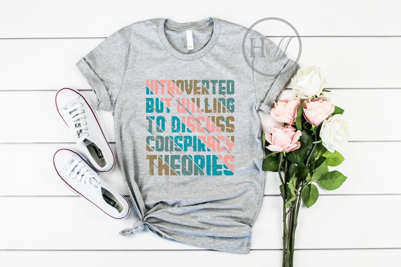 May include: Gray t-shirt with the text "Introverted but willing to discuss conspiracy theories" in a colorful retro font.
