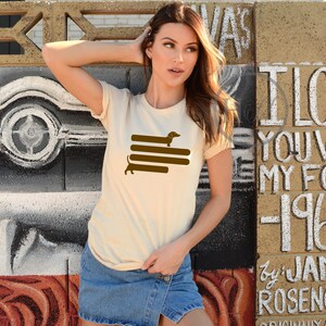 May include: A woman wearing a cream colored t-shirt with a brown dachshund silhouette graphic. The graphic shows the dachshund walking on top of four brown stripes. The woman is standing in front of a wall with white graffiti.