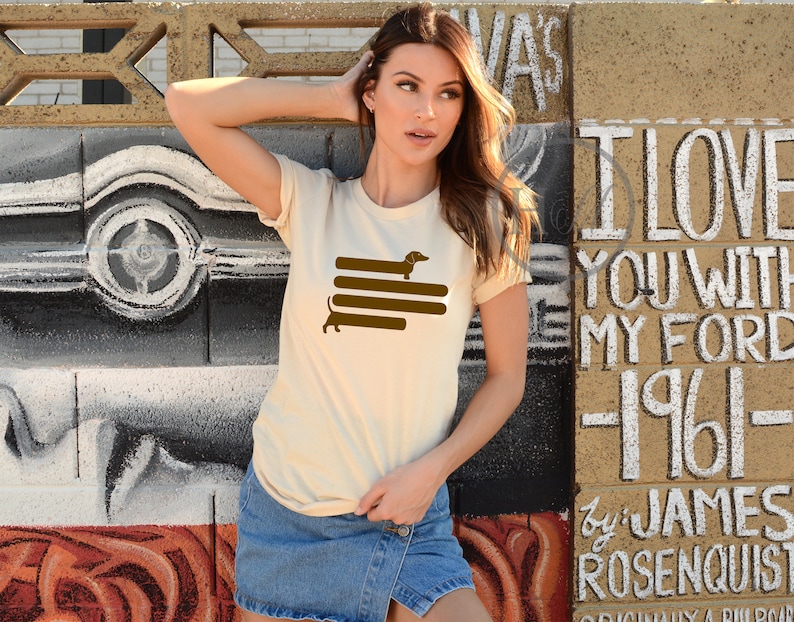 May include: A woman wearing a cream colored t-shirt with a brown dachshund silhouette graphic. The graphic shows the dachshund walking on top of four brown stripes. The woman is standing in front of a wall with white graffiti.