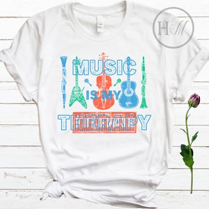 Pode incluir: Camiseta branca com o texto "Music is my therapy" impresso em azul, vermelho e verde. O texto &eacute; cercado por imagens de instrumentos musicais, incluindo uma guitarra, um violino, um clarinete e um teclado.