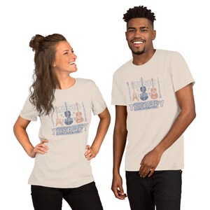 Pode incluir: Duas pessoas vestindo camisetas bege com um gr&aacute;fico de estilo vintage de instrumentos musicais e o texto "Music Therapy".