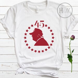 May include: White t-shirt featuring a red silhouette of a person wearing a hat, encircled by stars, with the number "45" above. The design is centered on the front of the shirt.