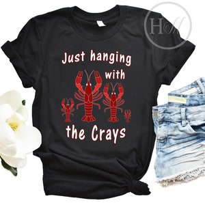 以下が含まれることがあります： 白と赤の文字で「Just hanging with the Crays」と書かれた黒いTシャツ。Tシャツには、赤いザリガニ3匹のグラフィックが描かれている。