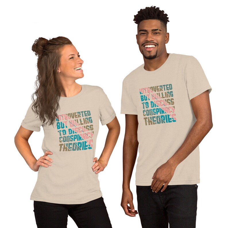 May include: Two people wearing beige t-shirts with the text "Introverted but willing to discuss conspiracy theories" printed in pink, blue, and teal. The text is stacked vertically and slanted.