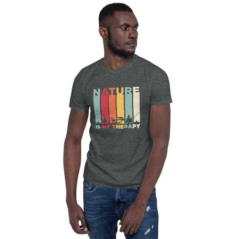 Puede incluir: Una camiseta gris oscuro jaspeada con un gr&aacute;fico de estilo vintage que dice "NATURE IS MY THERAPY". El gr&aacute;fico presenta un fondo a rayas de colores con una silueta de &aacute;rboles y un ciervo.