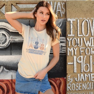 Pode incluir: Uma mulher vestindo uma camiseta bege com o texto "Music is my therapy" e um gr&aacute;fico de instrumentos musicais. A camiseta &eacute; combinada com uma saia jeans.