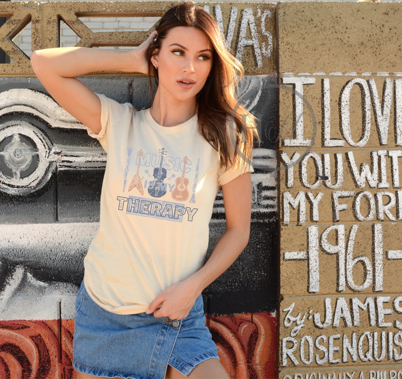Pode incluir: Uma mulher vestindo uma camiseta bege com o texto "Music is my therapy" e um gr&aacute;fico de instrumentos musicais. A camiseta &eacute; combinada com uma saia jeans.