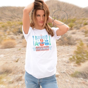 Pode incluir: Camiseta branca com um design gr&aacute;fico de instrumentos musicais e o texto "Music is my therapy".