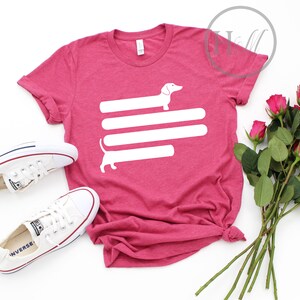 May include: A pink t-shirt with a white dachshund silhouette design. The dachshund is stretched out horizontally with its tail curled up. The dachshund is on top of three horizontal white lines.