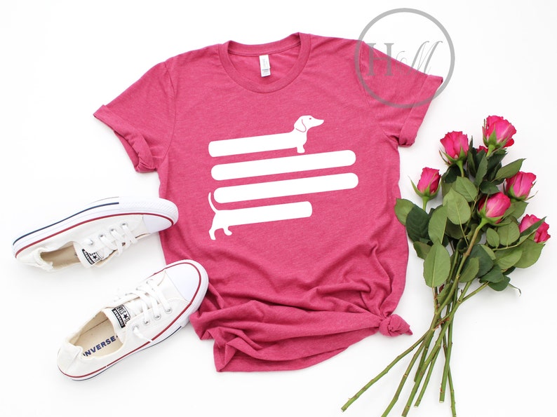 May include: A pink t-shirt with a white dachshund silhouette design. The dachshund is stretched out horizontally with its tail curled up. The dachshund is on top of three horizontal white lines.