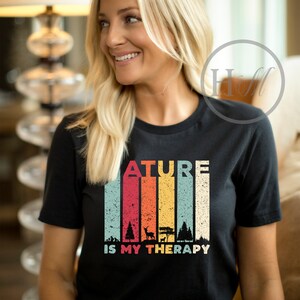 Puede incluir: Una camiseta negra con un gr&aacute;fico de estilo vintage que dice "Nature is my therapy". El gr&aacute;fico presenta un fondo a rayas de colores con siluetas de &aacute;rboles y un ciervo.