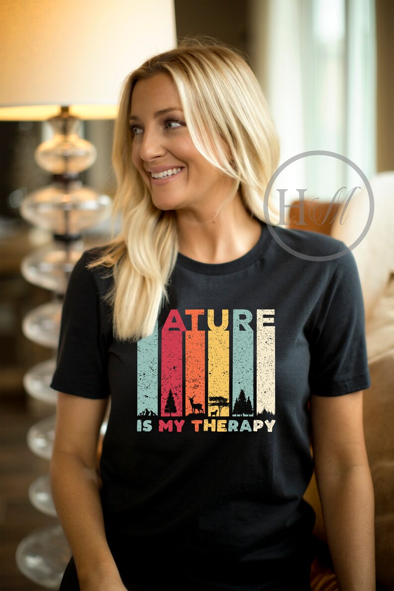 Puede incluir: Una camiseta negra con un gr&aacute;fico de estilo vintage que dice "Nature is my therapy". El gr&aacute;fico presenta un fondo a rayas de colores con siluetas de &aacute;rboles y un ciervo.