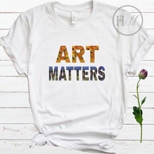 May include: White t-shirt with the text "ART MATTERS" in a stylized font. The letters are filled with a painting of sunflowers and a starry night.
