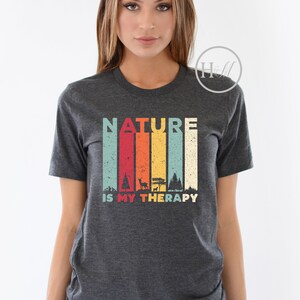 Puede incluir: Una camiseta gris oscuro jaspeada con un gr&aacute;fico de estilo vintage que dice "NATURE IS MY THERAPY". El gr&aacute;fico presenta una cordillera colorida con &aacute;rboles y un ciervo.