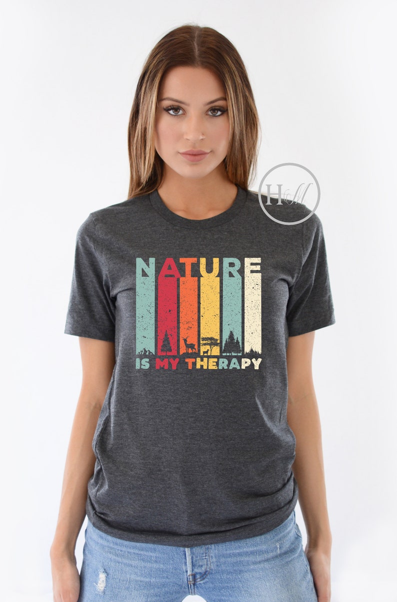 Puede incluir: Una camiseta gris oscuro jaspeada con un gr&aacute;fico de estilo vintage que dice "NATURE IS MY THERAPY". El gr&aacute;fico presenta una cordillera colorida con &aacute;rboles y un ciervo.