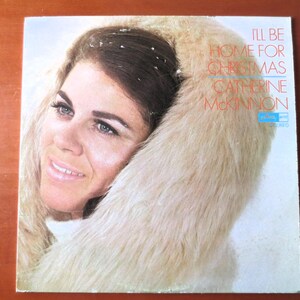 Puede incluir: Una portada de álbum de vinilo vintage con una mujer de cabello largo y castaño que lleva un abrigo de piel blanco. El título del álbum es "I'll Be Home For Christmas" de Catherine McKinnon.