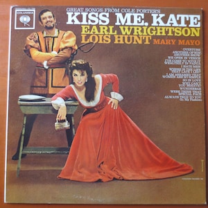 Puede incluir: Una portada de disco de vinilo vintage con una foto de dos actores disfrazados. La portada se titula "Kiss Me, Kate" con los nombres Earl Wrightson, Lois Hunt y Mary Mayo impresos debajo. La portada es de color marrón claro con un borde dorado. Los actores están vestidos con ropa de época renacentista. La mujer lleva un vestido rojo y el hombre lleva un traje amarillo y marrón. La portada incluye una lista de canciones del musical.