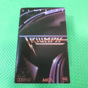 Puede incluir: Una caja de cassette con un dise&ntilde;o negro y plateado. La caja presenta el nombre de la banda "Triumph" en letras grandes y en negrita. El texto "Classics" est&aacute; impreso en letras m&aacute;s peque&ntilde;as en la parte superior de la caja. La caja tambi&eacute;n incluye los logotipos de MCA y HQ.