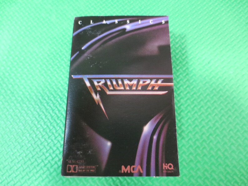 Puede incluir: Una caja de cassette con un dise&ntilde;o negro y plateado. La caja presenta el nombre de la banda "Triumph" en letras grandes y en negrita. El texto "Classics" est&aacute; impreso en letras m&aacute;s peque&ntilde;as en la parte superior de la caja. La caja tambi&eacute;n incluye los logotipos de MCA y HQ.