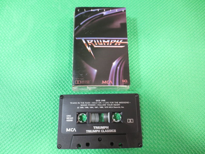 Puede incluir: Una cinta de casete negra con el t&iacute;tulo "Triumph Classics" y el nombre de la banda "Triumph" en letras blancas. La cinta de casete tiene una caja negra con una ventana de pl&aacute;stico transparente que muestra la cinta en el interior. La cinta de casete est&aacute; etiquetada con los t&iacute;tulos de las canciones "Tears in the Rain", "Hold On", "Live for the Weekend", "Magic Power" y "Follow Your Heart".