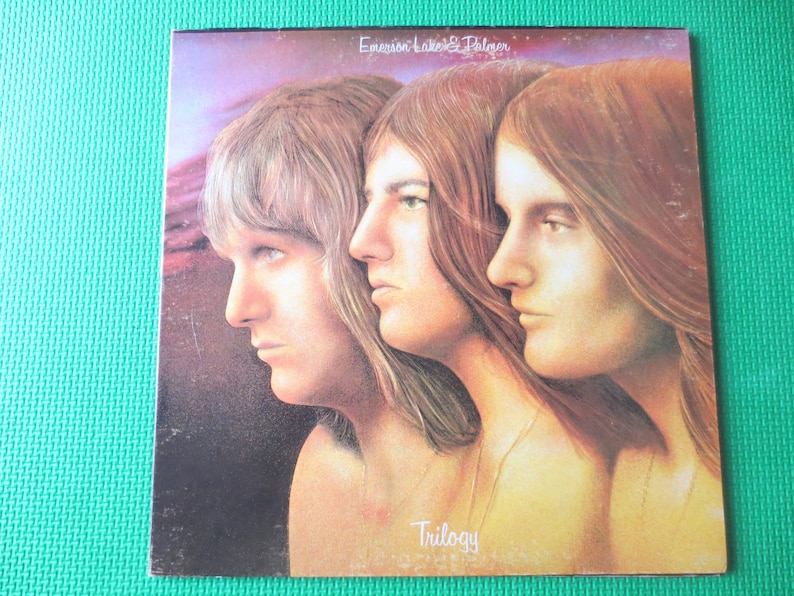 May include: A vintage vinyl record album cover featuring a photo of three men with long hair. The album is titled "Trilogy" by Emerson, Lake & Palmer.