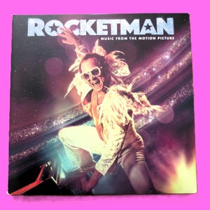 May include: Album cover for the film "Rocketman" showing a person in a white outfit, sunglasses, and feathered wings. The title is in silver letters with a star. The background is a starry, purple and pink space.