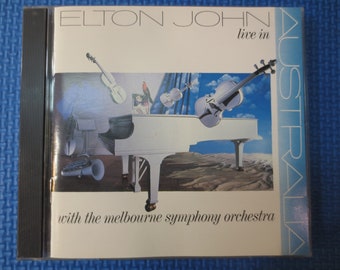 May include: A white CD case with a photo of a white grand piano and three violins. The text on the case reads "Elton John live in Australia with the Melbourne Symphony Orchestra".