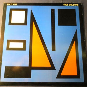 Może przedstawiać: Okładka winylowego albumu w kolorze niebieskim z tytułem "True Colours" i nazwą artysty "Split Enz" w białym kolorze. Okładka zawiera geometryczny wzór z czarnymi, białymi i pomarańczowymi kształtami.