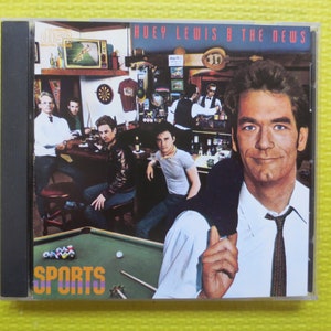 May include: A CD case with a photo of Huey Lewis and the News on the cover. The band is in a bar setting with a pool table in the foreground. The album title "Sports" is printed in large, colorful letters at the bottom of the cover.