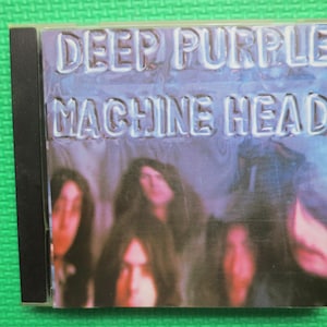 May include: A CD case with a purple and blue background featuring the text "Deep Purple Machine Head". The case has a black spine and shows a blurry image of four people.