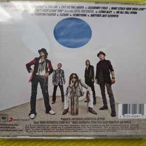 May include: A CD case with a white background and a blue circle in the center. The case features a group of five people standing in front of a white wall. The text on the case reads "Aerosmith - Tell Me, Out Go The Lights, Legendary Child, What Could Have Been Love, Can't Stop Lovin' You, Lover Alot, We All Fall Down, Freedom Fighter, Closer, Something, Another Last Goodbye".