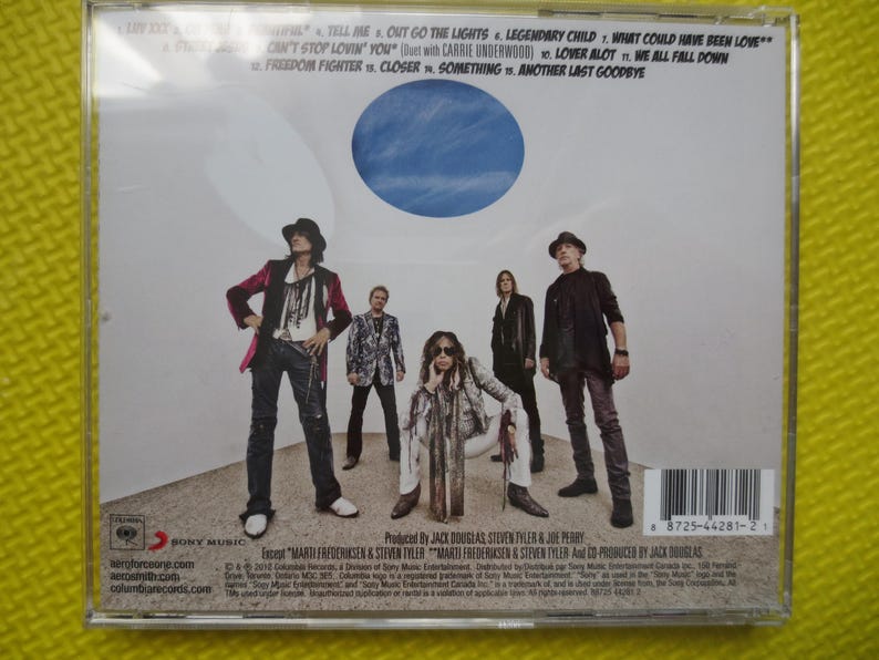 May include: A CD case with a white background and a blue circle in the center. The case features a group of five people standing in front of a white wall. The text on the case reads "Aerosmith - Tell Me, Out Go The Lights, Legendary Child, What Could Have Been Love, Can't Stop Lovin' You, Lover Alot, We All Fall Down, Freedom Fighter, Closer, Something, Another Last Goodbye".
