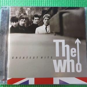 Puede incluir: Una caja de CD plateada con una foto en blanco y negro de los miembros de la banda The Who. El texto "The Who" y "Greatest Hits" est&aacute; impreso en la parte delantera de la caja.