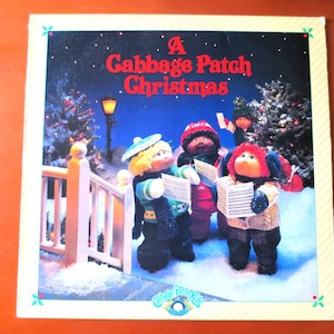 Könnte beinhalten: Ein Plattencover von Cabbage Patch Kids Christmas mit vier Puppen, die vor einer verschneiten Winterlandschaft mit einem Weihnachtsbaum und einer Straßenlaterne Weihnachtslieder singen.