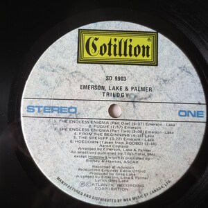 May include: A white vinyl record with black text. The record is labeled "Cotillion" and "SD 9903". The text "Emerson, Lake & Palmer" and "Trilogy" is also printed on the record. The record is labeled "Stereo" and "One".