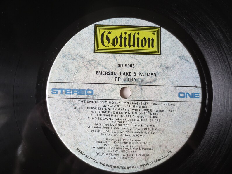 May include: A white vinyl record with black text. The record is labeled "Cotillion" and "SD 9903". The text "Emerson, Lake & Palmer" and "Trilogy" is also printed on the record. The record is labeled "Stereo" and "One".