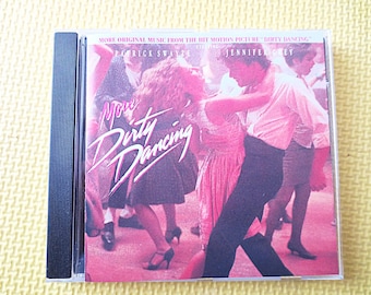May include: A pink and white CD case with the title "More Dirty Dancing" in pink and white lettering. The cover features a photo of Patrick Swayze and Jennifer Grey dancing.