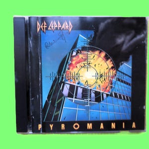 Könnte beinhalten: Ein Def Leppard "Pyromania" CD-Album. Das Cover zeigt ein Fadenkreuz über einem Gebäude mit einer feurigen Explosion. Das Logo der Band befindet sich oben und der Albumtitel unten. Die CD-Hülle ist schwarz.