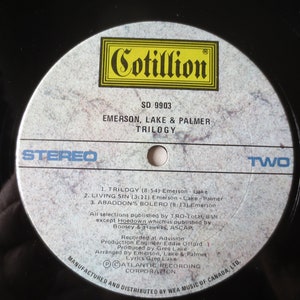 May include: A white vinyl record with a black label. The label has the text "Cotillion" in a yellow box, "SD 9903", "Emerson, Lake & Palmer", "Trilogy", "Stereo", and "Two". The record lists the song titles "Trilogy", "Living Sin", and "Abaddon's Bolero".