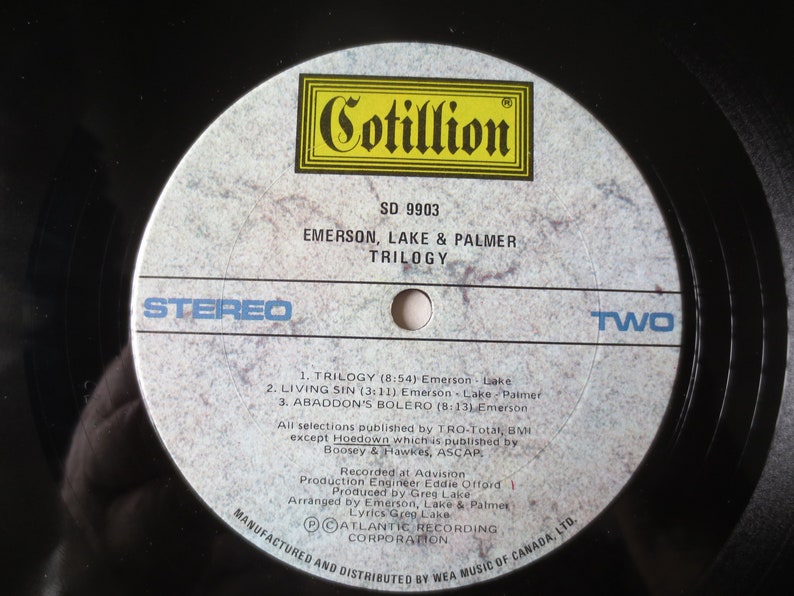 May include: A white vinyl record with a black label. The label has the text "Cotillion" in a yellow box, "SD 9903", "Emerson, Lake & Palmer", "Trilogy", "Stereo", and "Two". The record lists the song titles "Trilogy", "Living Sin", and "Abaddon's Bolero".
