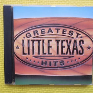 May include: A CD case with a brown wooden background. The text "Greatest Little Texas Hits" is in a black oval with white stars on either side.