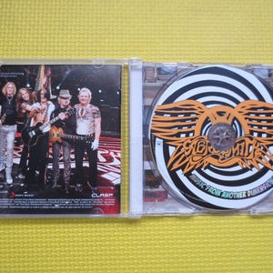 May include: A CD case with a black and white photo of a rock band on the front cover. The back cover features a band logo and the text "Music from Another Dimension!" The CD case is open, revealing a CD with a black and white spiral design and the band's logo in orange.