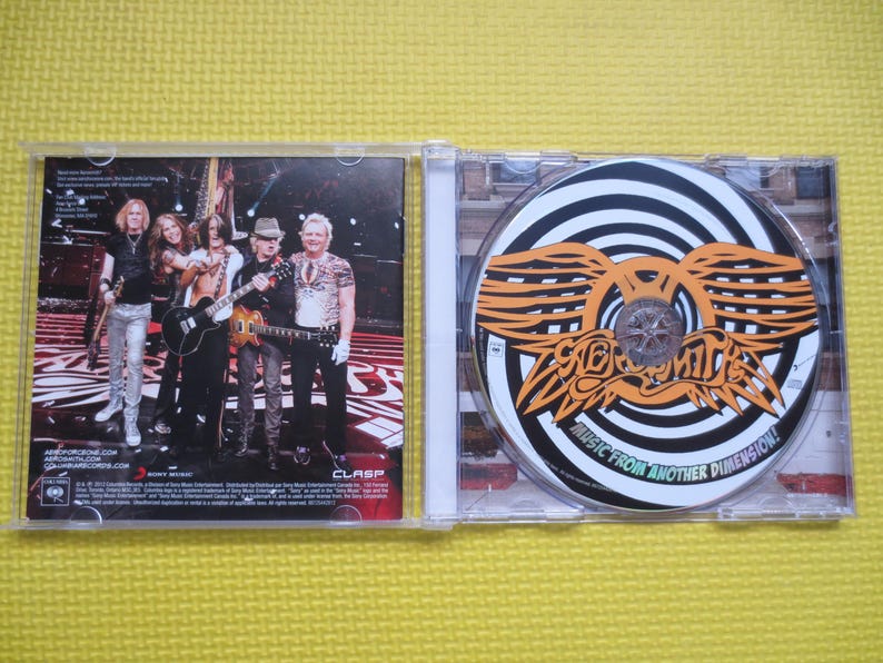 May include: A CD case with a black and white photo of a rock band on the front cover. The back cover features a band logo and the text "Music from Another Dimension!" The CD case is open, revealing a CD with a black and white spiral design and the band's logo in orange.
