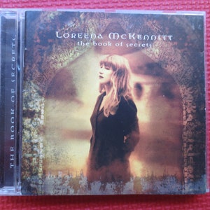 Könnte beinhalten: Das Albumcover von Loreena McKennitt für "The Book of Secrets". Das Cover zeigt eine Frau mit langen braunen Haaren, die in einem Tor mit einem Steinbogen steht. Der Hintergrund ist ein verblasstes, sepiafarbenes Bild.