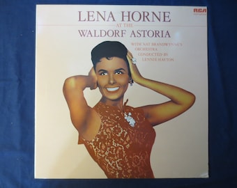 Pode incluir: Uma capa de álbum de vinil vintage com Lena Horne, uma famosa cantora e atriz, com um sorriso caloroso. O álbum é intitulado "Lena Horne at the Waldorf Astoria" com a orquestra de Nat Brandwynne conduzida por Lennie Hayton.