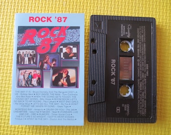 May include: A black cassette tape with a white label that says "Rock '87". The cassette tape has a picture of a group of people on the front. The back of the cassette tape has a list of songs on it.